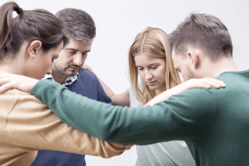 Photo of four people in a huddle with eyes closed and hands on each others shoulders, offering support to one another